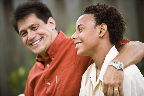 An older man with his arm loosely around a teenager's shoulder in a friendly manner. They are both smiling.